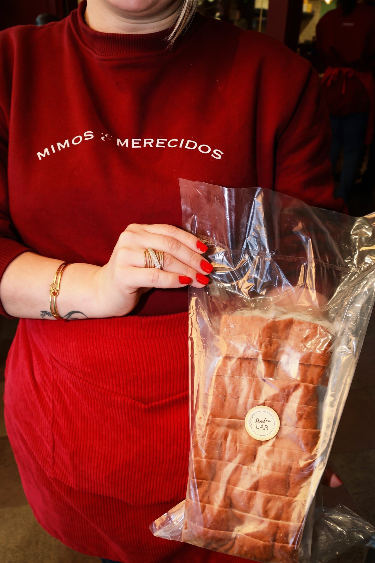 Pessoa de suéter vermelho bordado com “Mimos & merecidos” segura um pão de forma fatiado no saquinho.