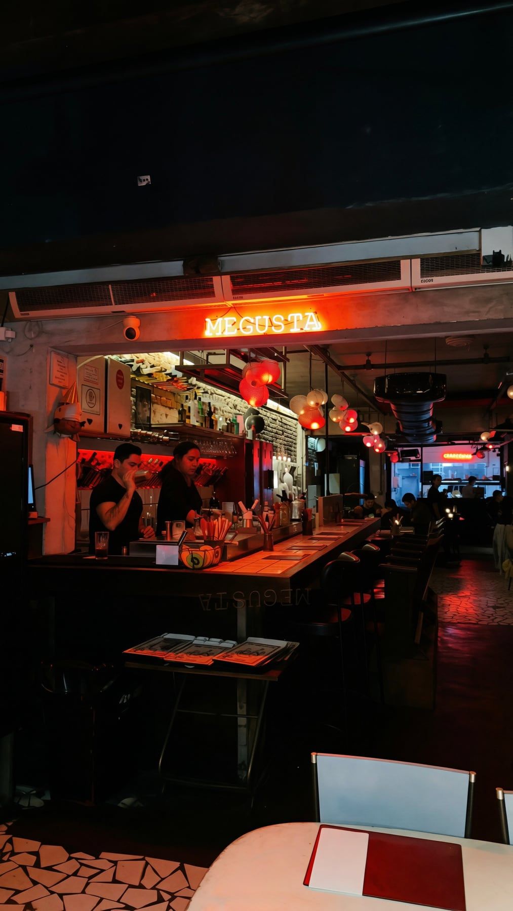 O balcão do MeGusta à noite: letreiro vermelho com o nome da casa ao fundo, bartenders trabalhando e banquetas ao longo do bar.
