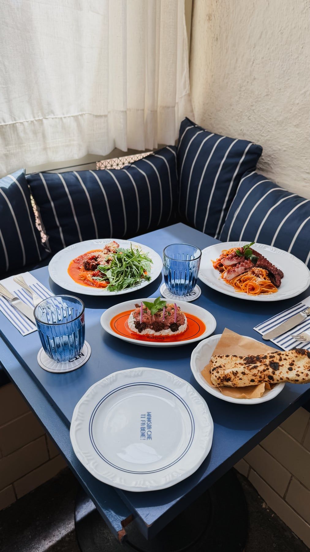 Banco estofado com almofadas listradas de azul e branco junto a uma mesa azul servida com pratos e copos azuis.