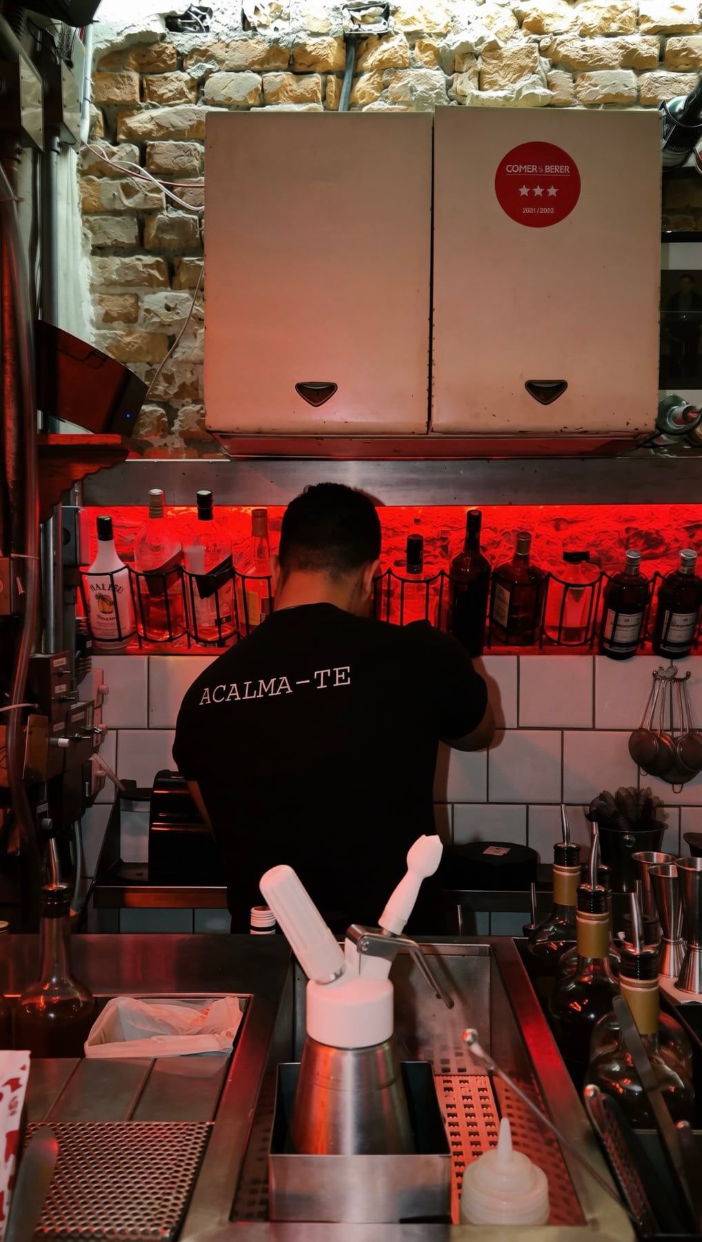 Bartender de costas, de camiseta escrita Acalma-te, preparando drinks sob luz vermelha, diante de uma parede de pedra.