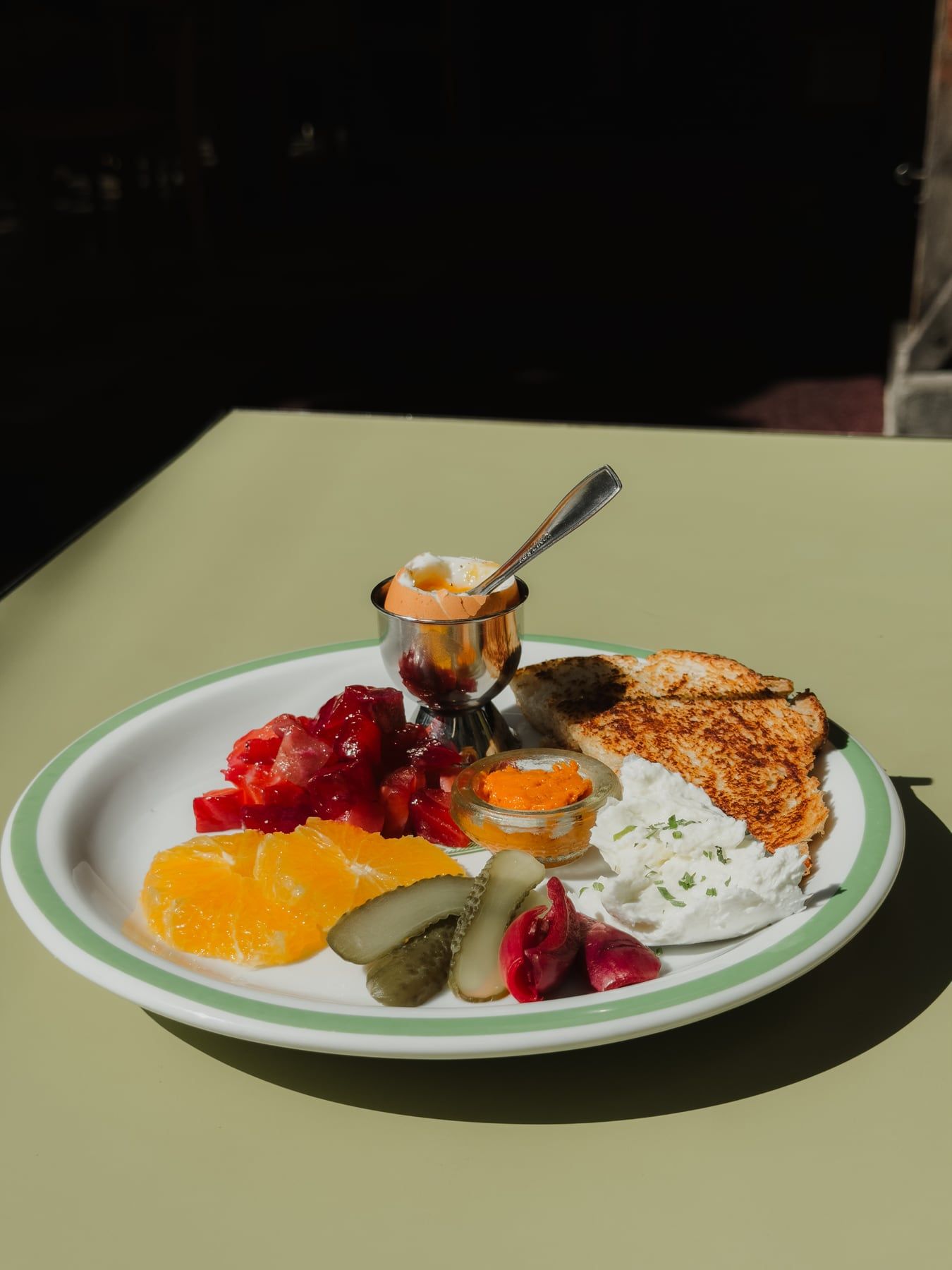 Prato de café da manhã ao sol: frutas, picles, creme com ervas, torrada e uma tacinha de inox.