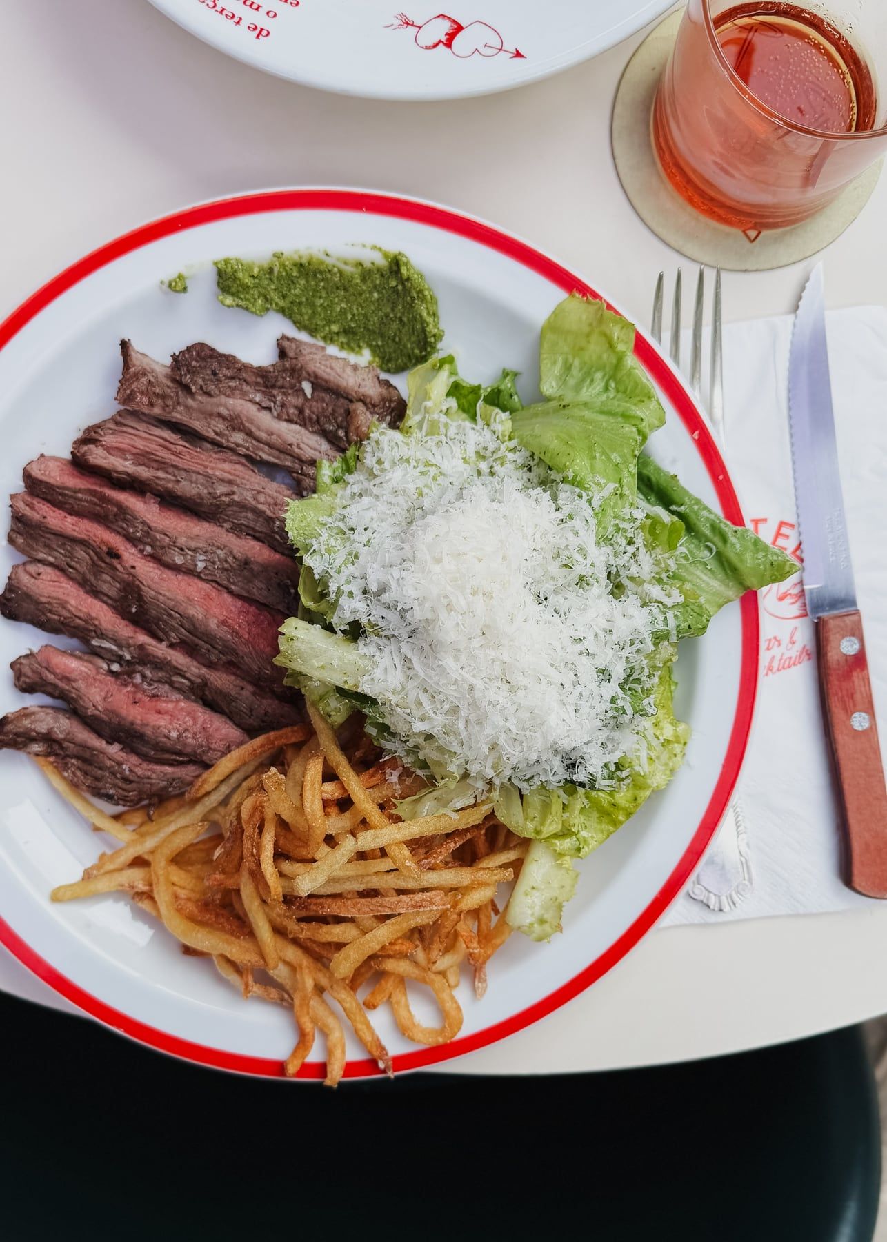 Carne fatiada ao ponto com batata palha, alface com queijo ralado e molho verde, em prato de borda vermelha.