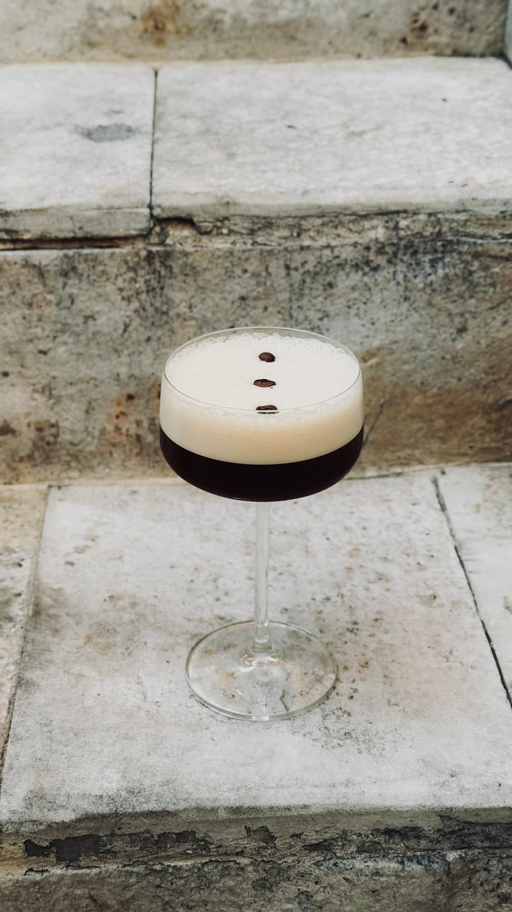 Drinque escuro numa taça, com espuma clara e três grãos de café por cima, sobre degraus de pedra.