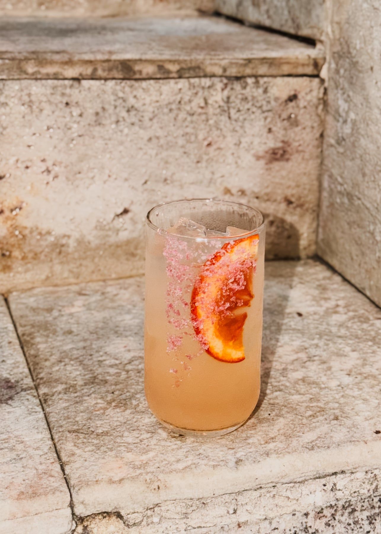 Drinque claro num copo alto com gelo, açúcar rosa na borda e uma rodela de laranja.
