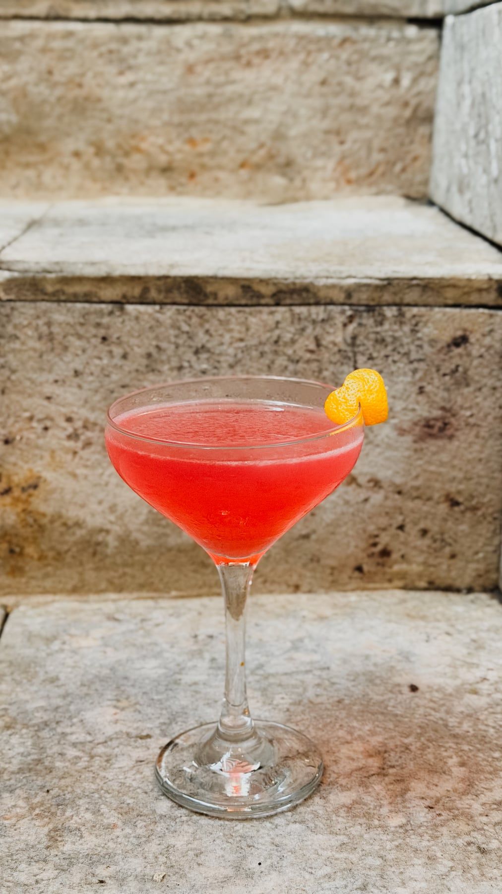 Drinque rosado numa taça de haste, com uma casca de laranja enrolada na borda.