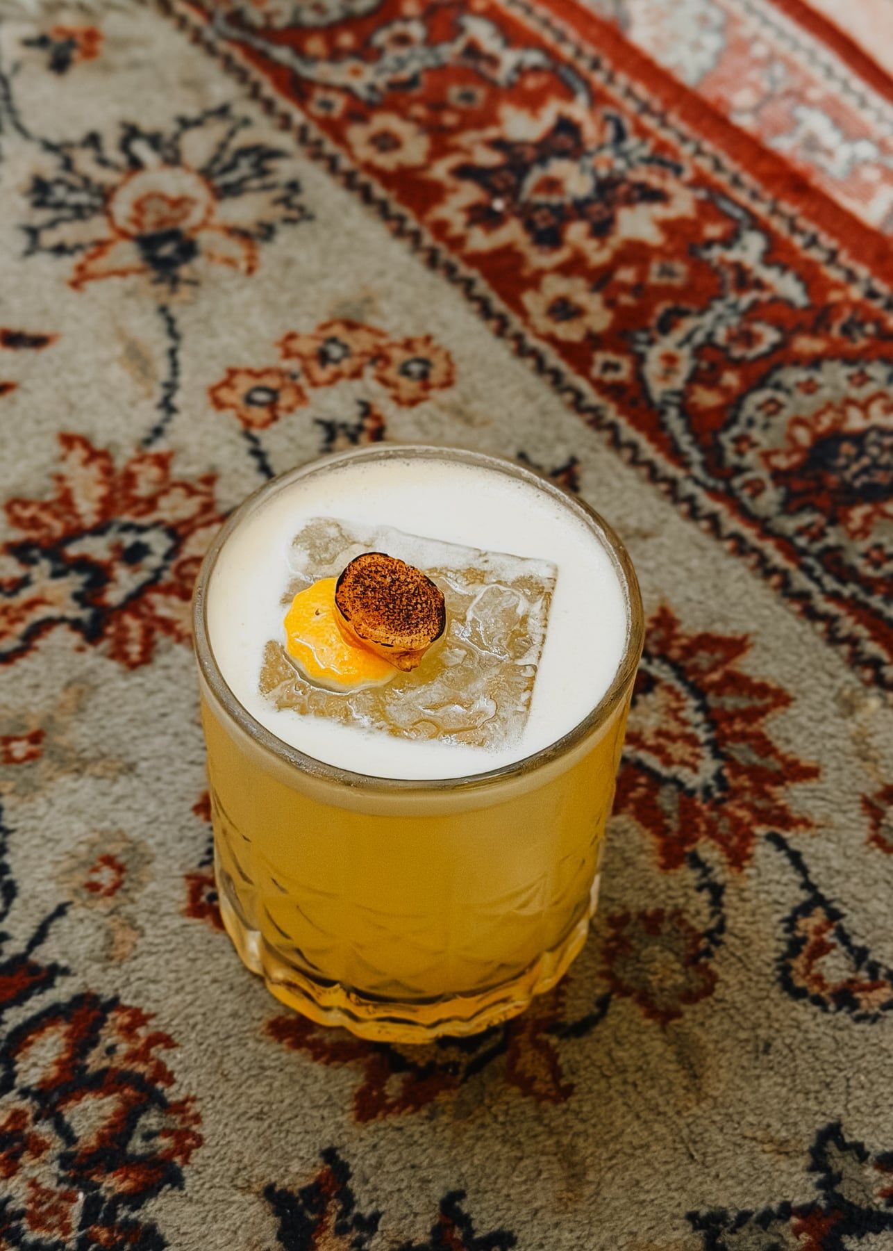 Drinque amarelo com espuma num copo de vidro lapidado, enfeitado com uma rodela tostada, sobre o tapete persa.