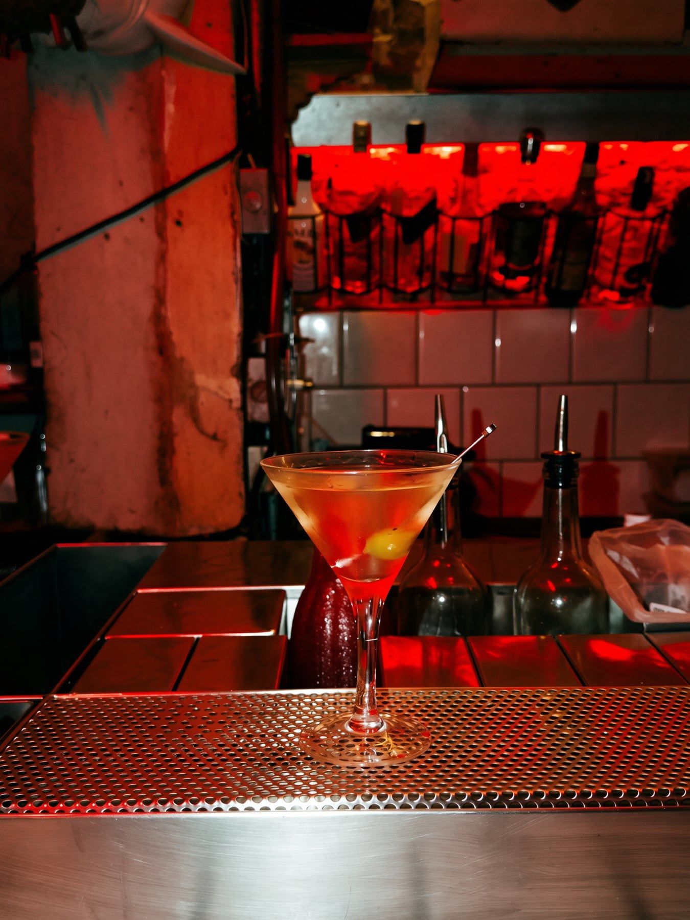 Dry martini com azeitona sobre o balcão de metal, com garrafas iluminadas de vermelho ao fundo.