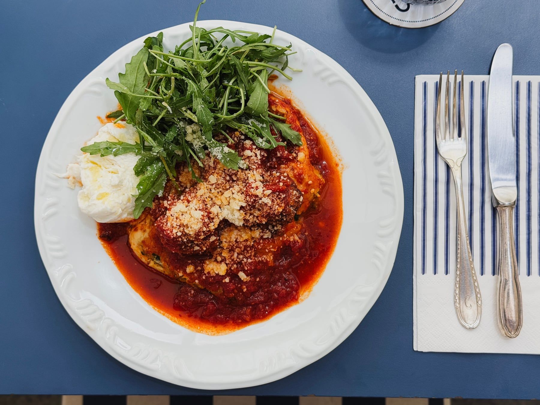 Massa gratinada com molho de tomate, rúcula e burrata, num prato branco de borda trabalhada.