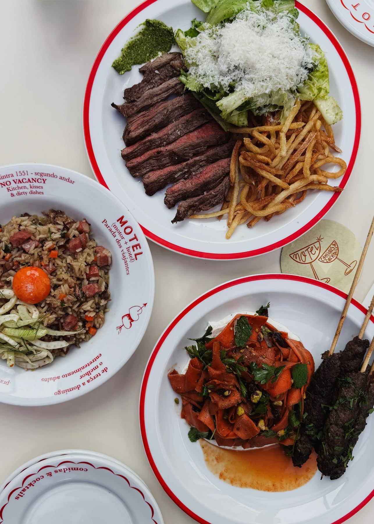 Mesa vista de cima com quatro pratos de borda vermelha para dividir: carne fatiada com fritas e salada, arroz com gema, cenoura com espetinhos de carne e outro prato com o nome Motel.