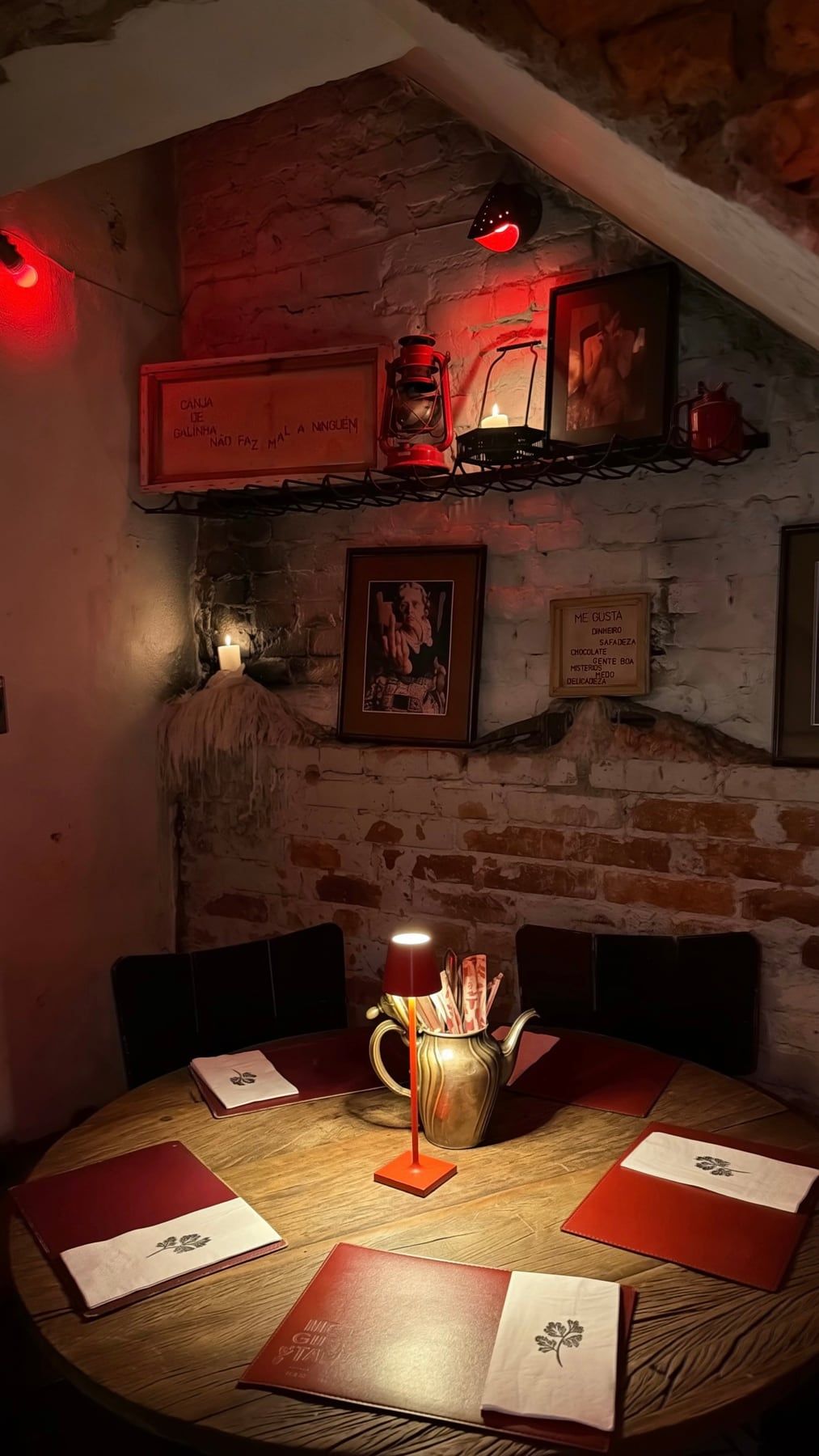 Mesa redonda de madeira à luz de um abajur, com cardápios, sob uma prateleira com lampião e parede de tijolos. Foto do MeGusta, usada aqui até o Mé ter fotos próprias.