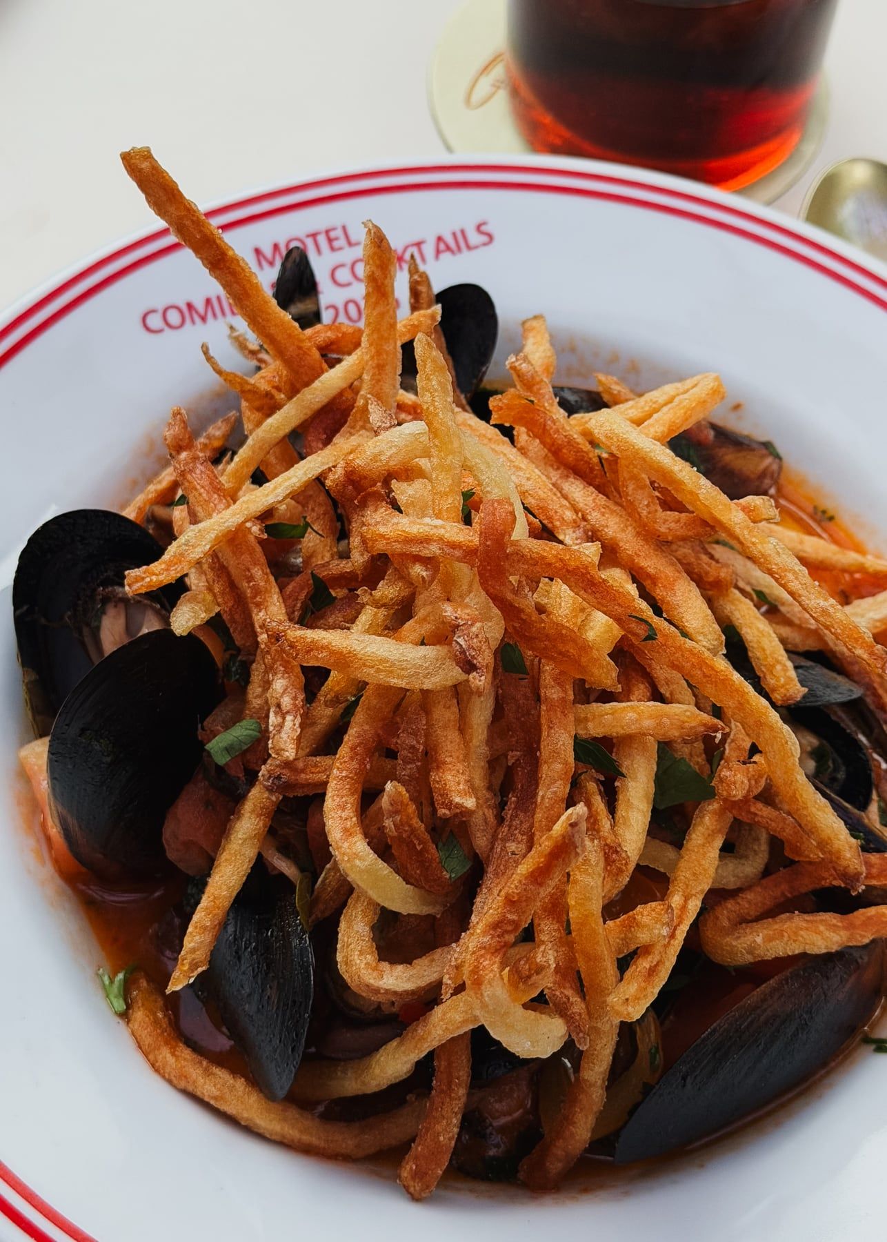 Mexilhões em molho de tomate cobertos por batata palha, num prato fundo com o nome Motel na borda.