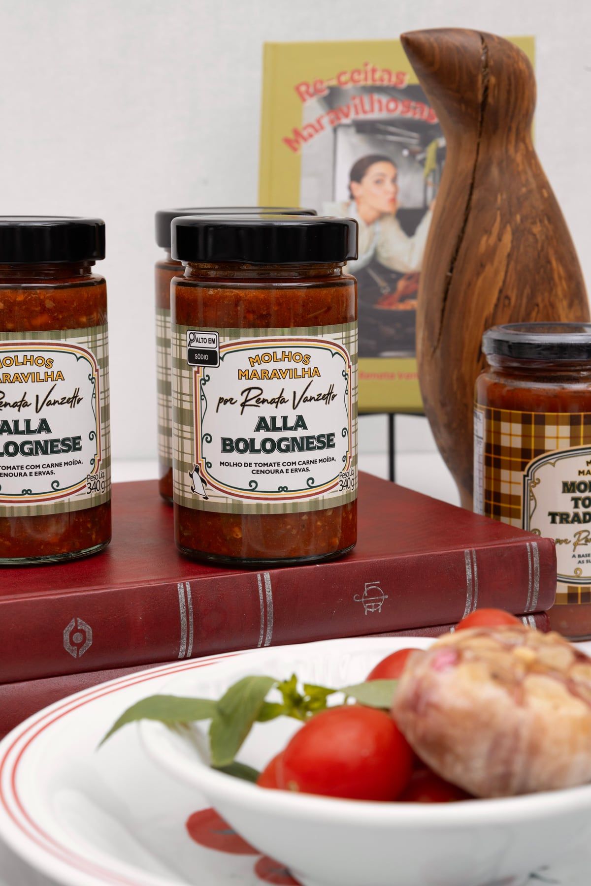 Potes de molho alla bolognese com rótulo xadrez “Molhos Maravilha por Renata Vanzetto”, sobre livros vermelhos, ao lado de um pinguim de madeira.