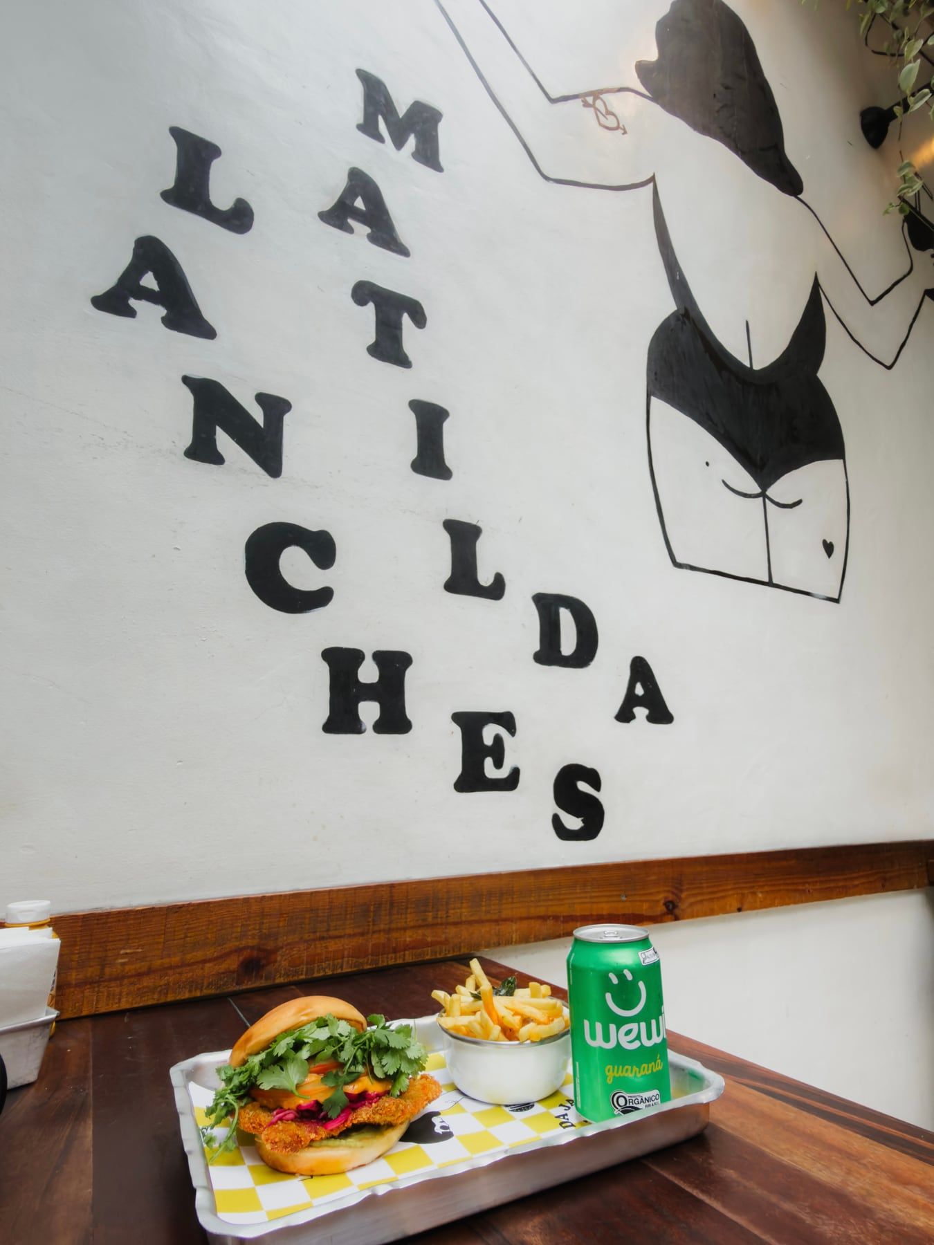Bandeja com hambúrguer, batata frita e guaraná no balcão de madeira, embaixo do letreiro Matilda Lanches pintado na parede.