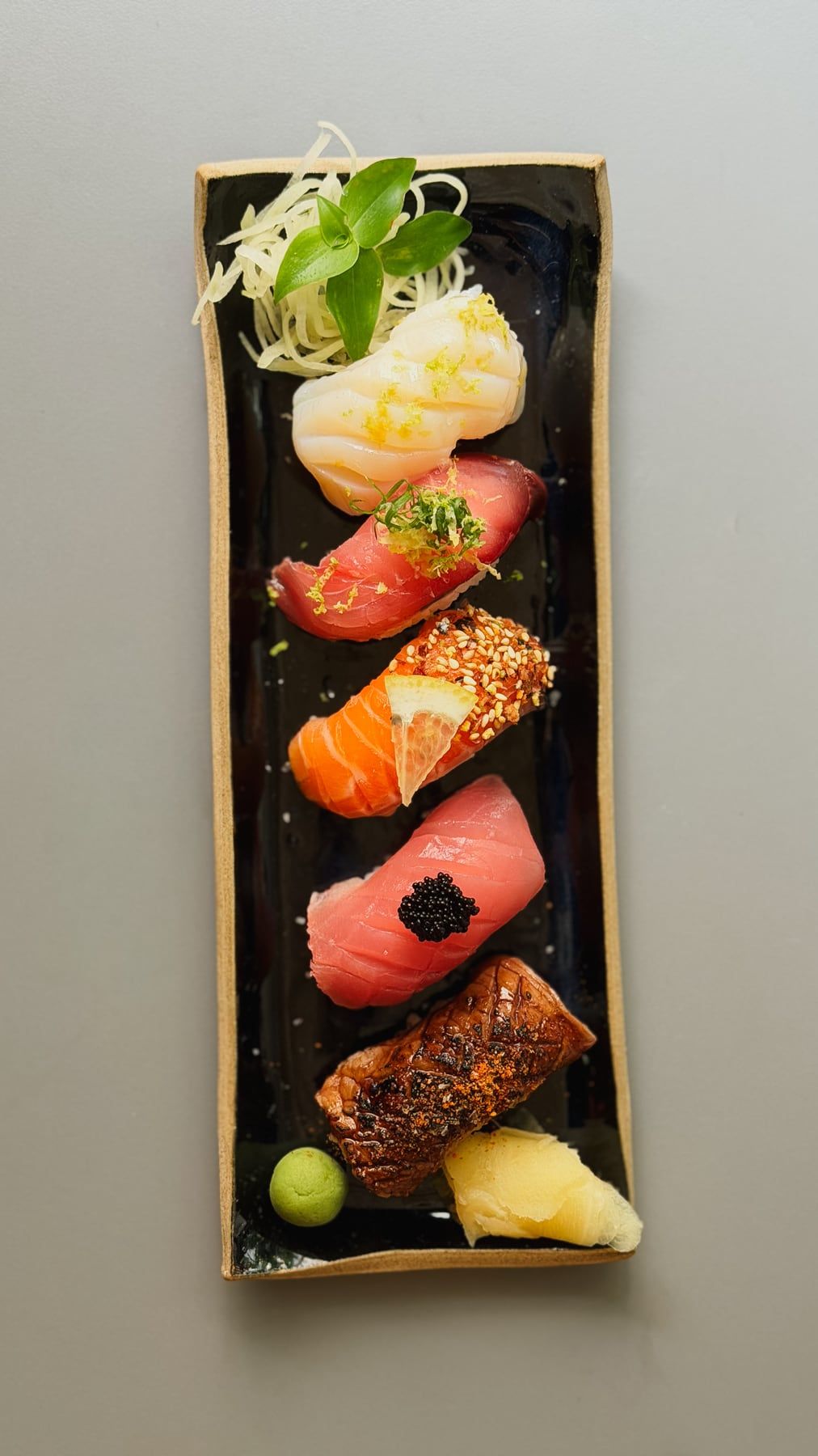 Travessa comprida de pedra escura com nigiris de vieira, atum, salmão e peixe maçaricado, com gengibre e wasabi.