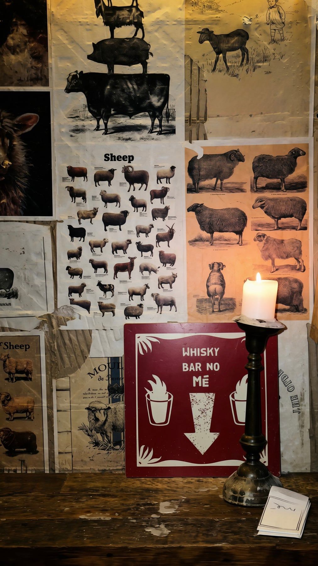 Placa vermelha escrita Whisky bar no Mé, com uma seta apontando para baixo, ao lado de uma vela acesa e cartazes antigos de ovelhas, no MeGusta.
