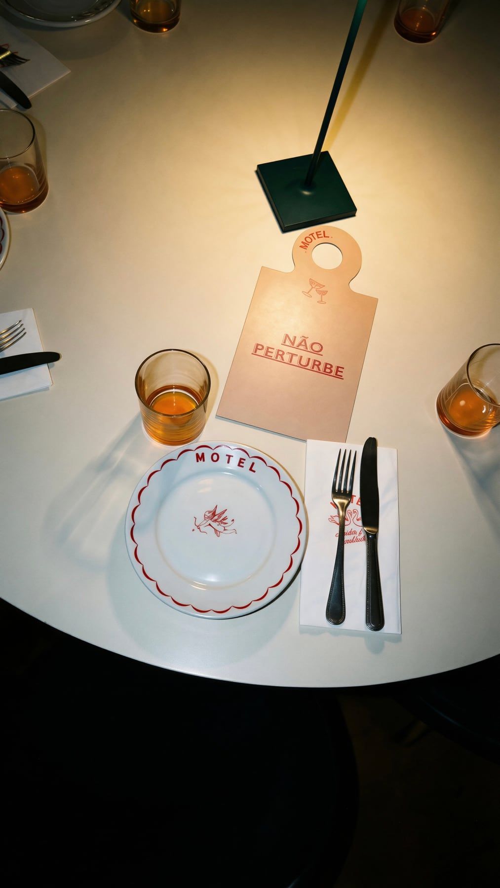 Mesa redonda branca com a plaquinha de porta escrita Não perturbe, um prato do Motel, talheres e dois copos de bebida âmbar.