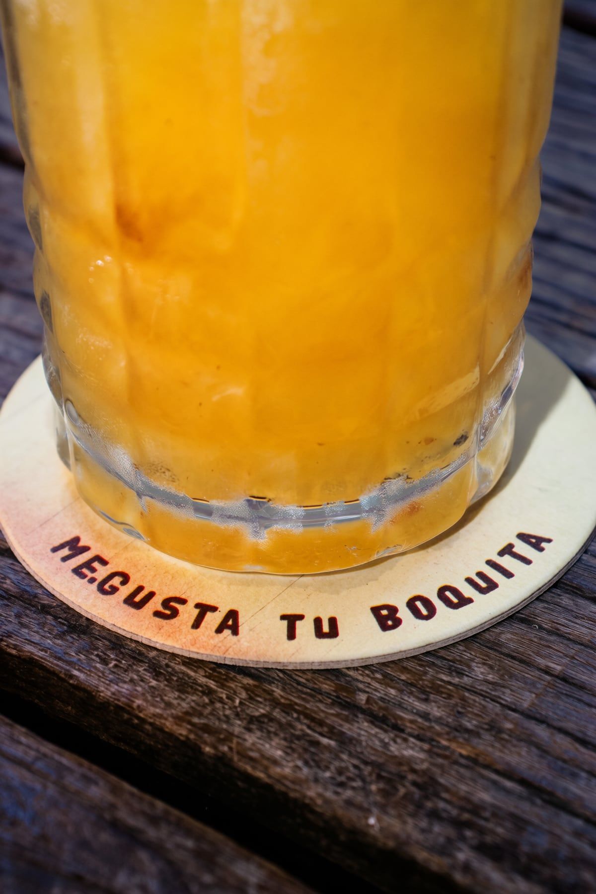 Copo de drink alaranjado sobre um porta-copo de papelão escrito Megusta tu boquita, numa mesa de madeira.