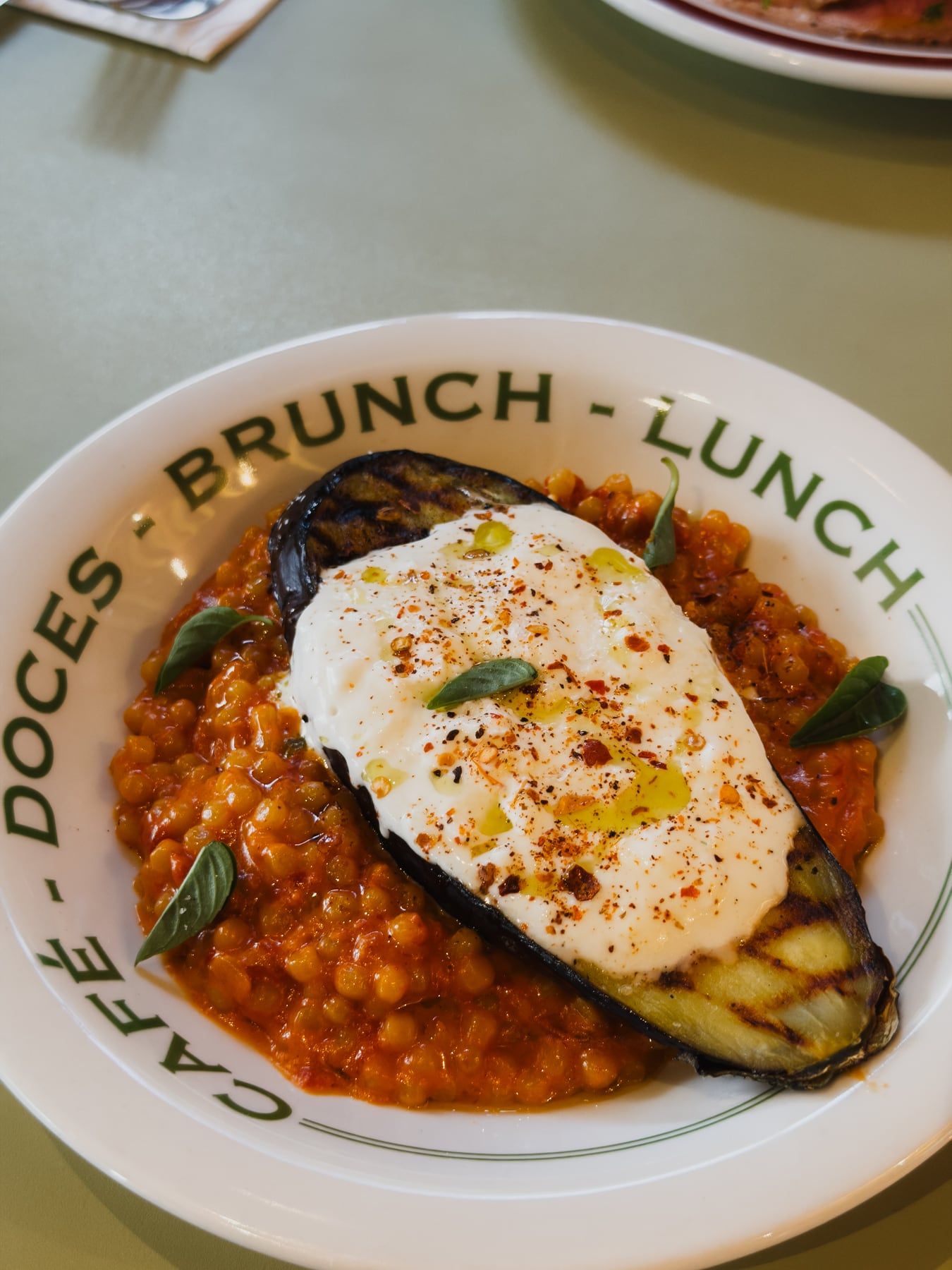 Prato de brunch na louça com a borda escrita “Café – Doces – Brunch – Lunch”: berinjela grelhada com creme branco, páprica e manjericão sobre grãos ao molho de tomate.
