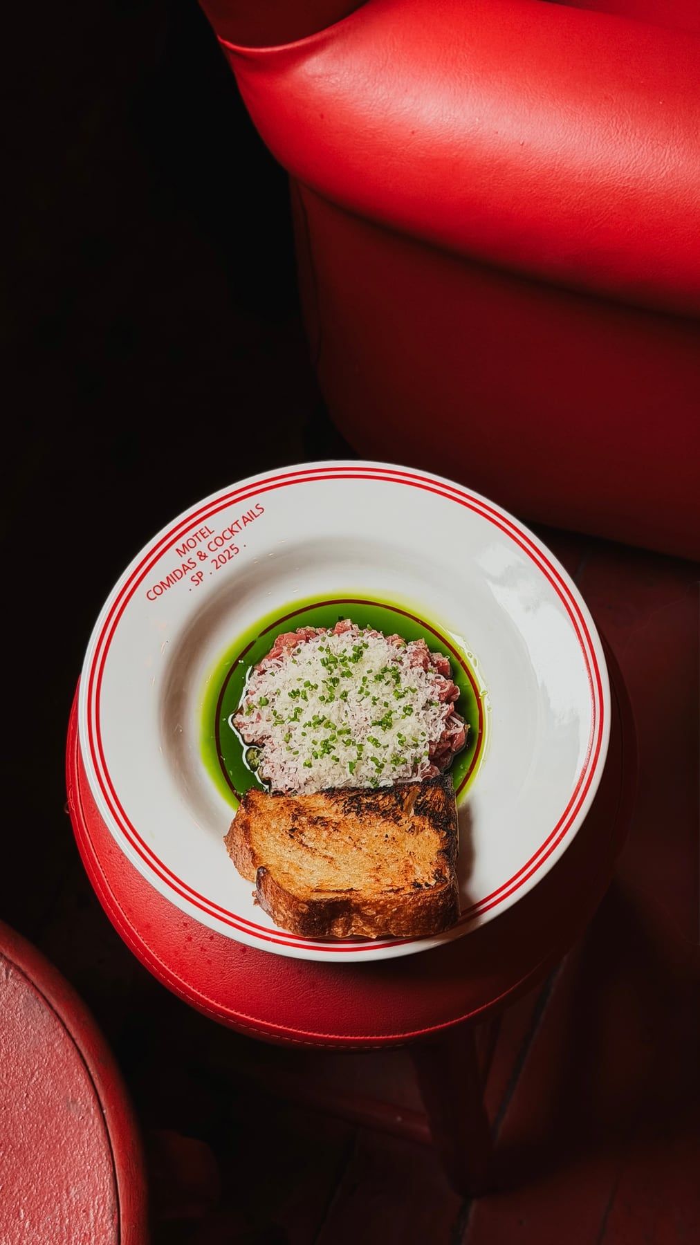 Prato raso escrito Motel, comidas e cocktails, com uma entrada e uma fatia de pão tostado, sobre uma mesa vermelha ao lado do banco de vinil.