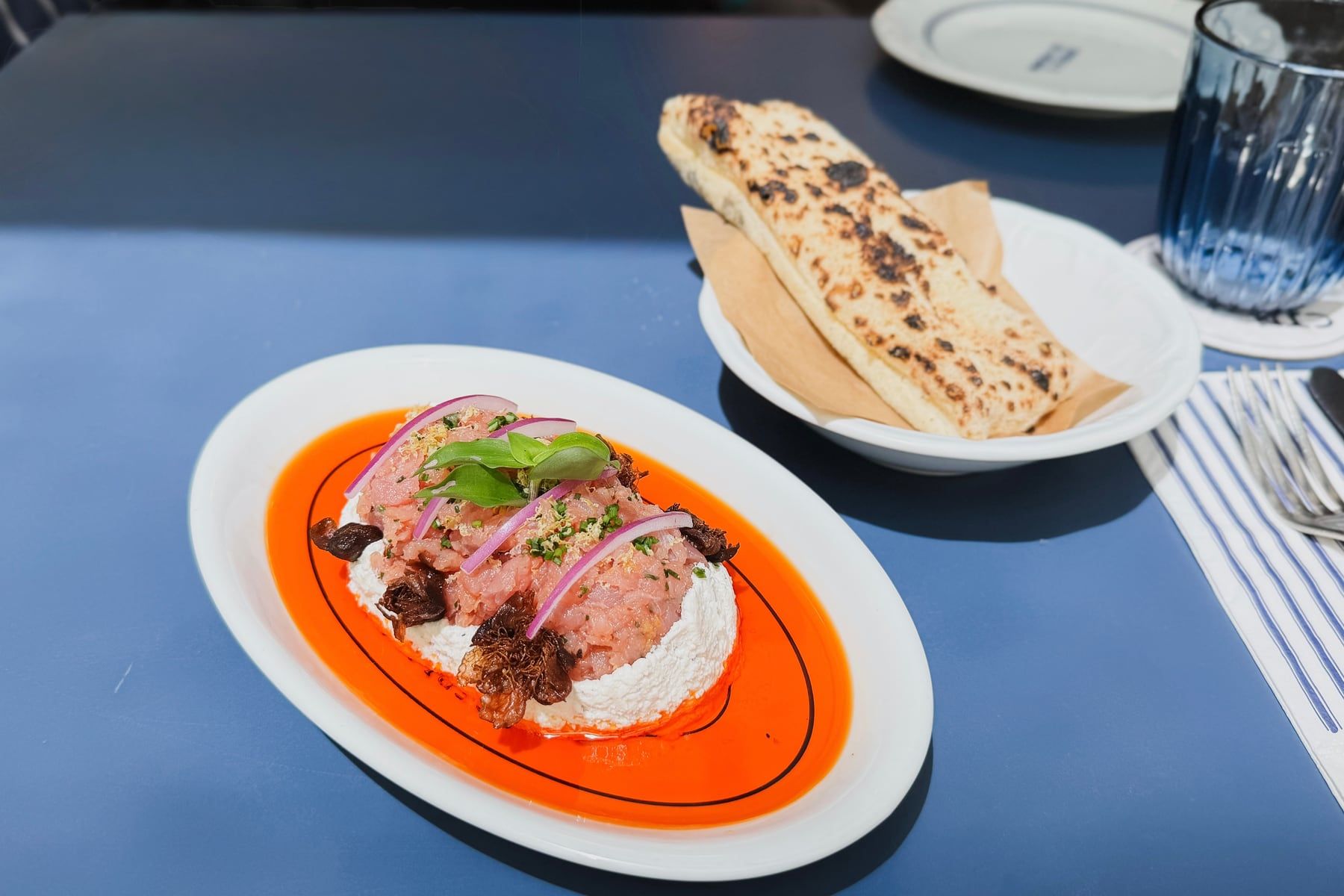De perto, o prato oval de borda laranja da casa, com carne crua picada, cebola roxa e manjericão sobre creme branco.