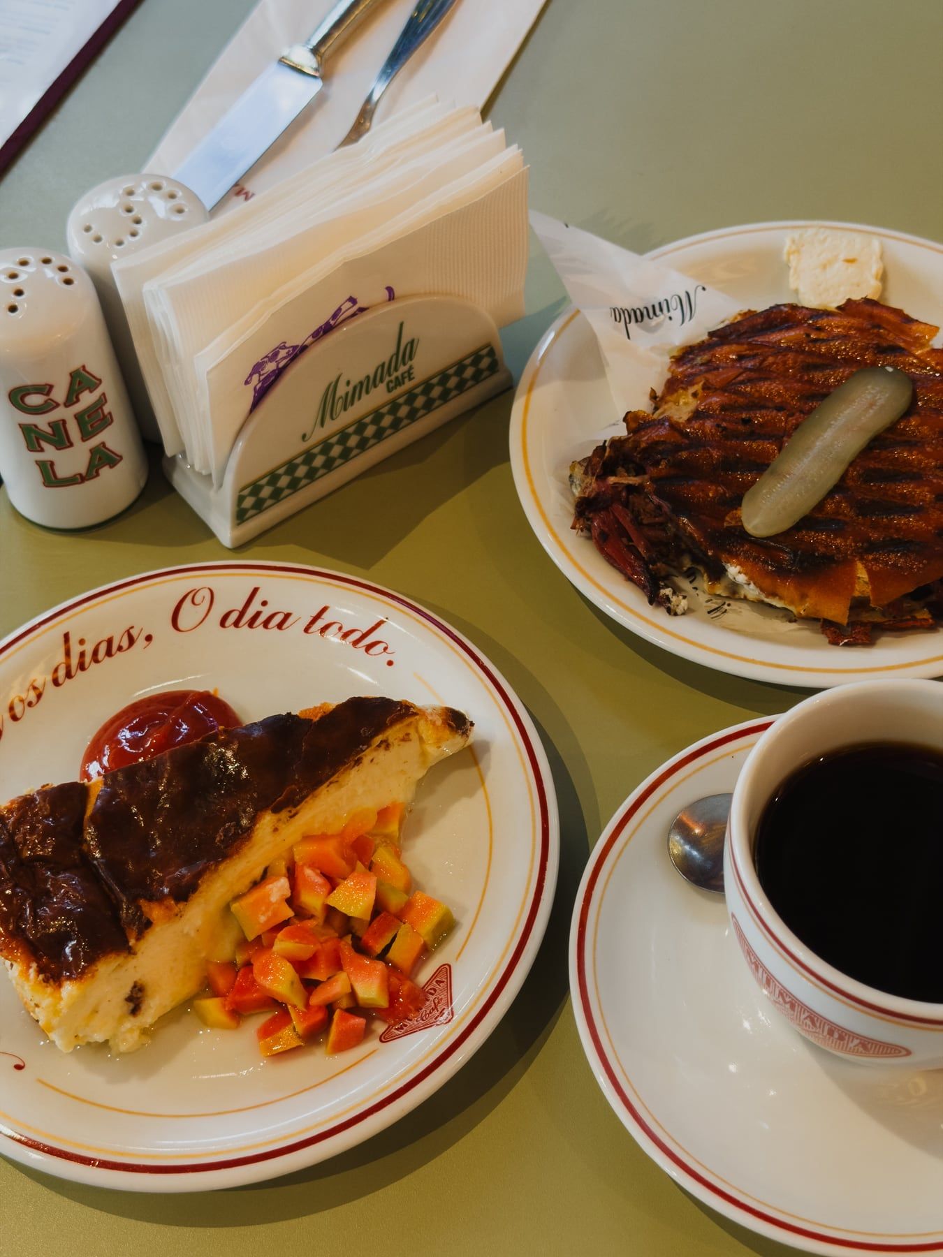 Café na mesa, visto de cima: fatia assada com calda escura e cubos de mamão no prato escrito “Todos os dias, o dia todo”, sanduíche na chapa e café coado.