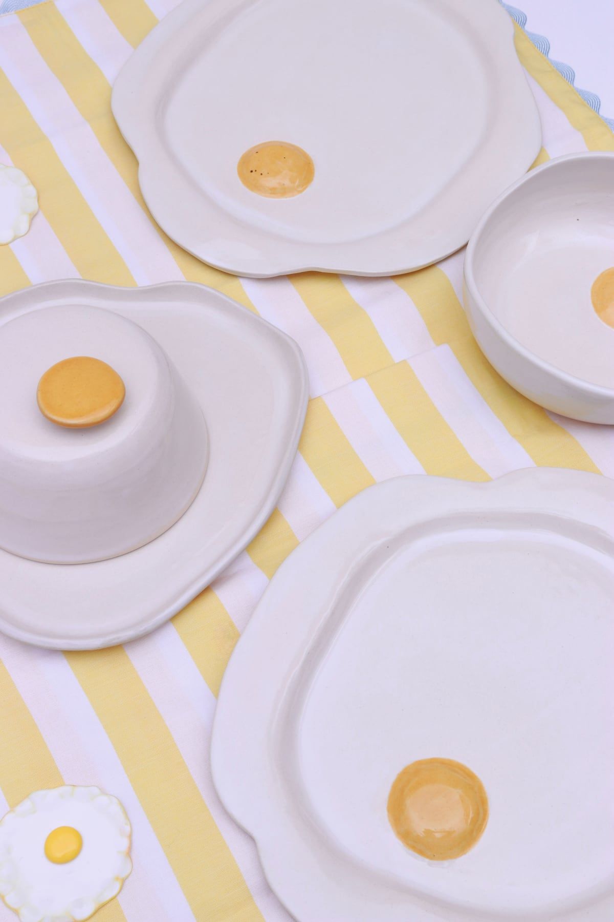 Pratos, tigela e queijeira de cerâmica branca em formato de ovo frito, com gemas amarelas, sobre uma toalha listrada de amarelo e branco.