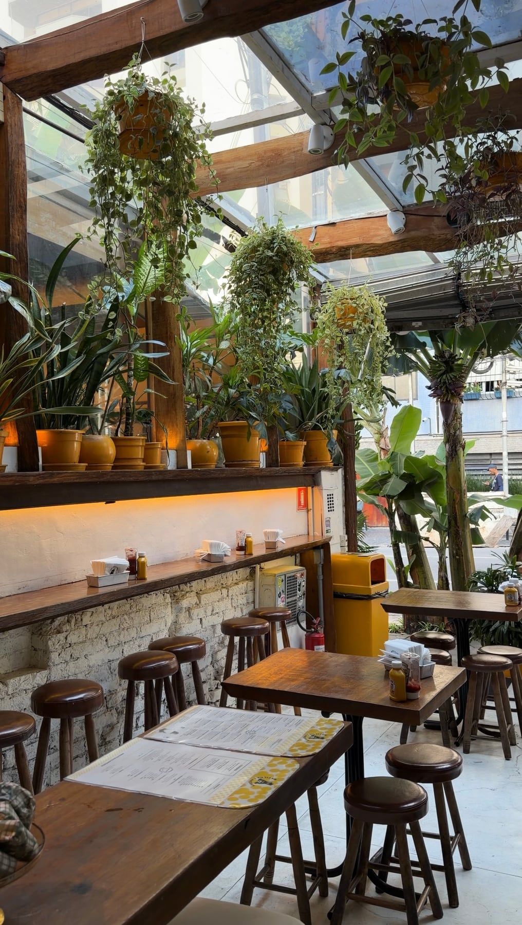 Salão com teto de vidro, vigas de madeira, vasos amarelos e muitas plantas sobre o balcão, mesas e banquetas de madeira.