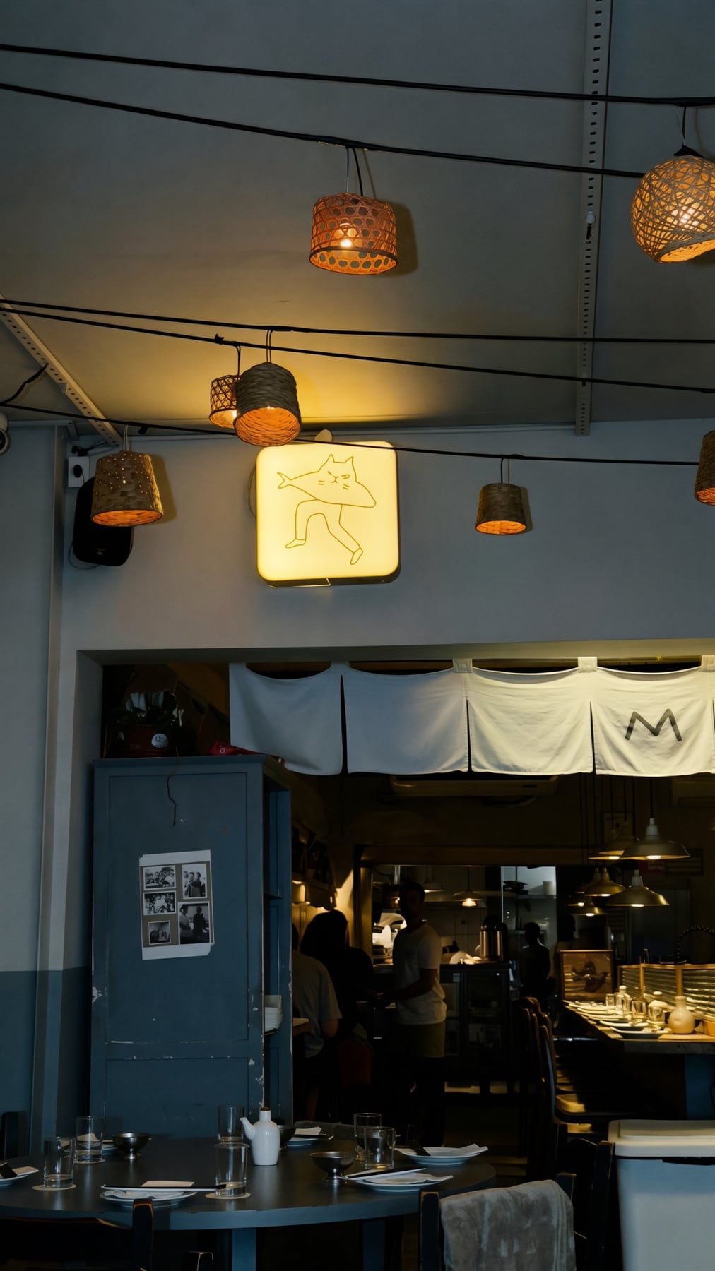Salão à meia-luz: luminárias de palha, o luminoso com o homem-gato-peixe e as cortinas de pano na entrada da cozinha.