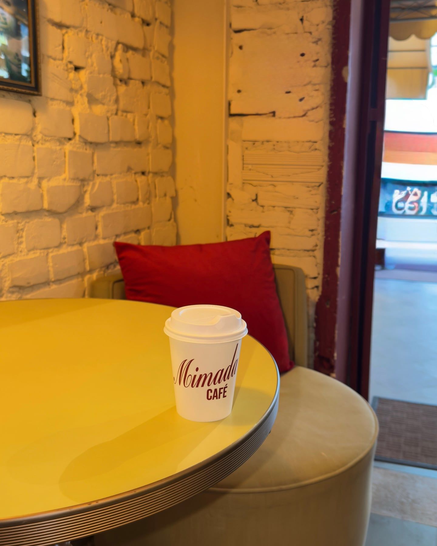 Canto do salão: mesa redonda amarela, almofada vermelha e um copo de café para viagem do Mimada, junto à parede de tijolos pintados.