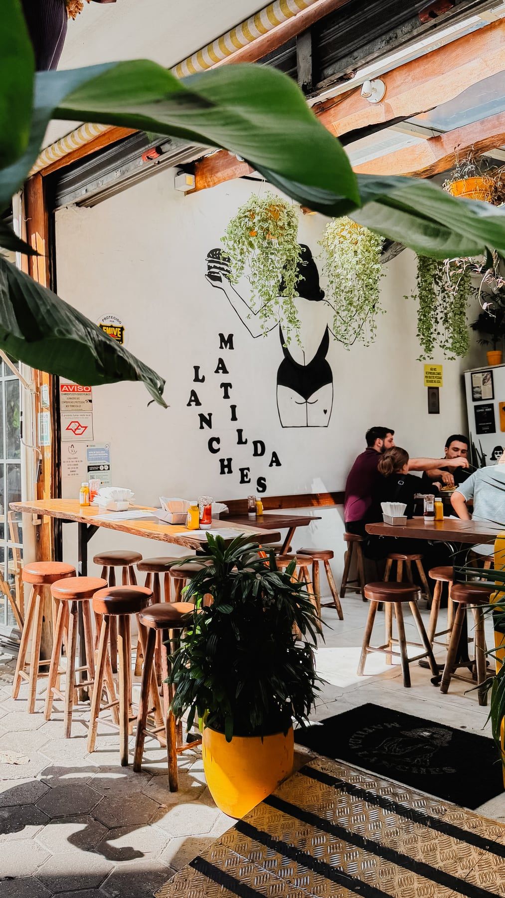 Salão com banquetas de madeira, plantas penduradas e, na parede branca, a Matilda pintada em preto segurando um hambúrguer, com as letras de Matilda Lanches espalhadas.