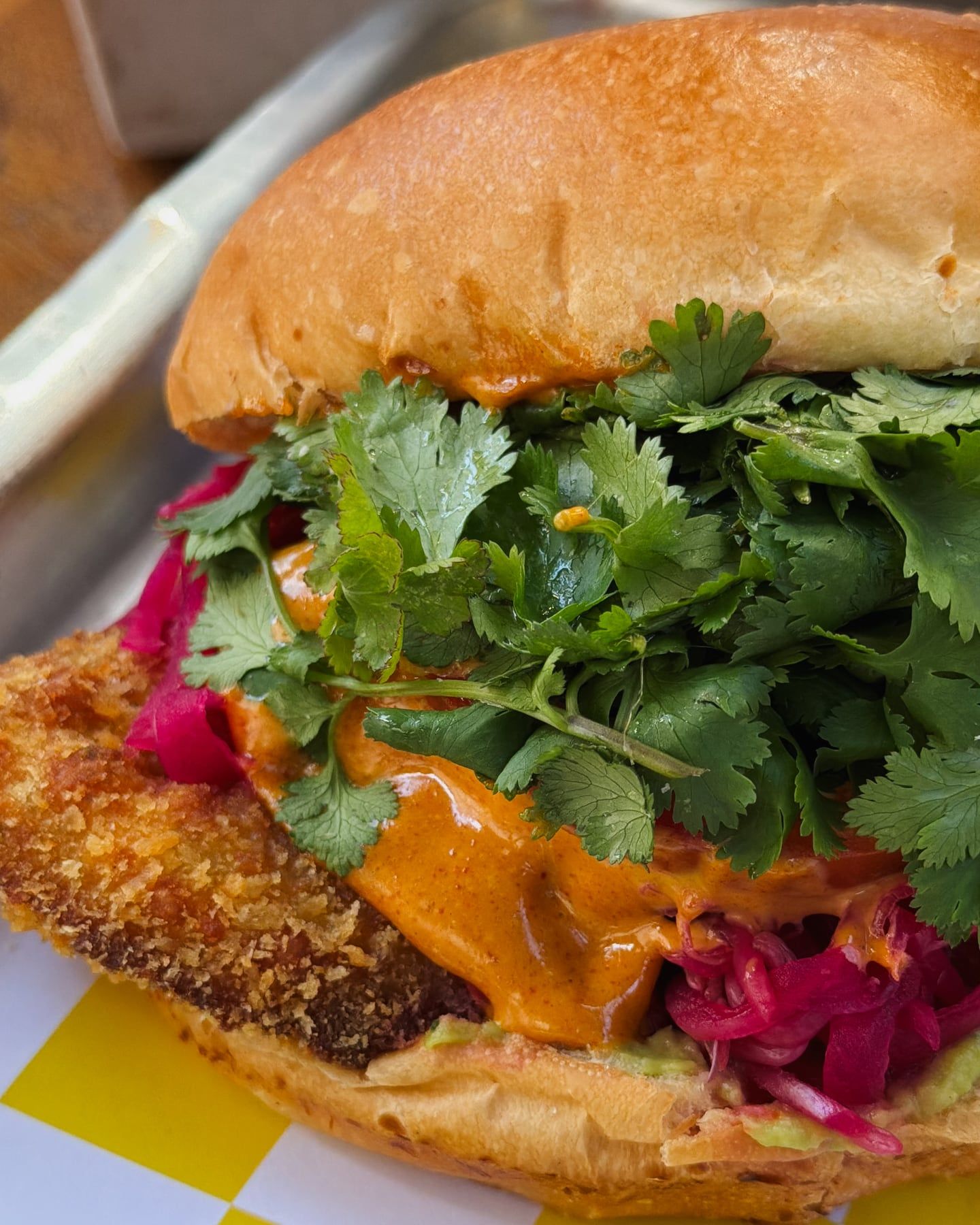 Sanduíche de perto: pão brioche, empanado crocante, molho alaranjado, coentro fresco e cebola roxa em conserva.