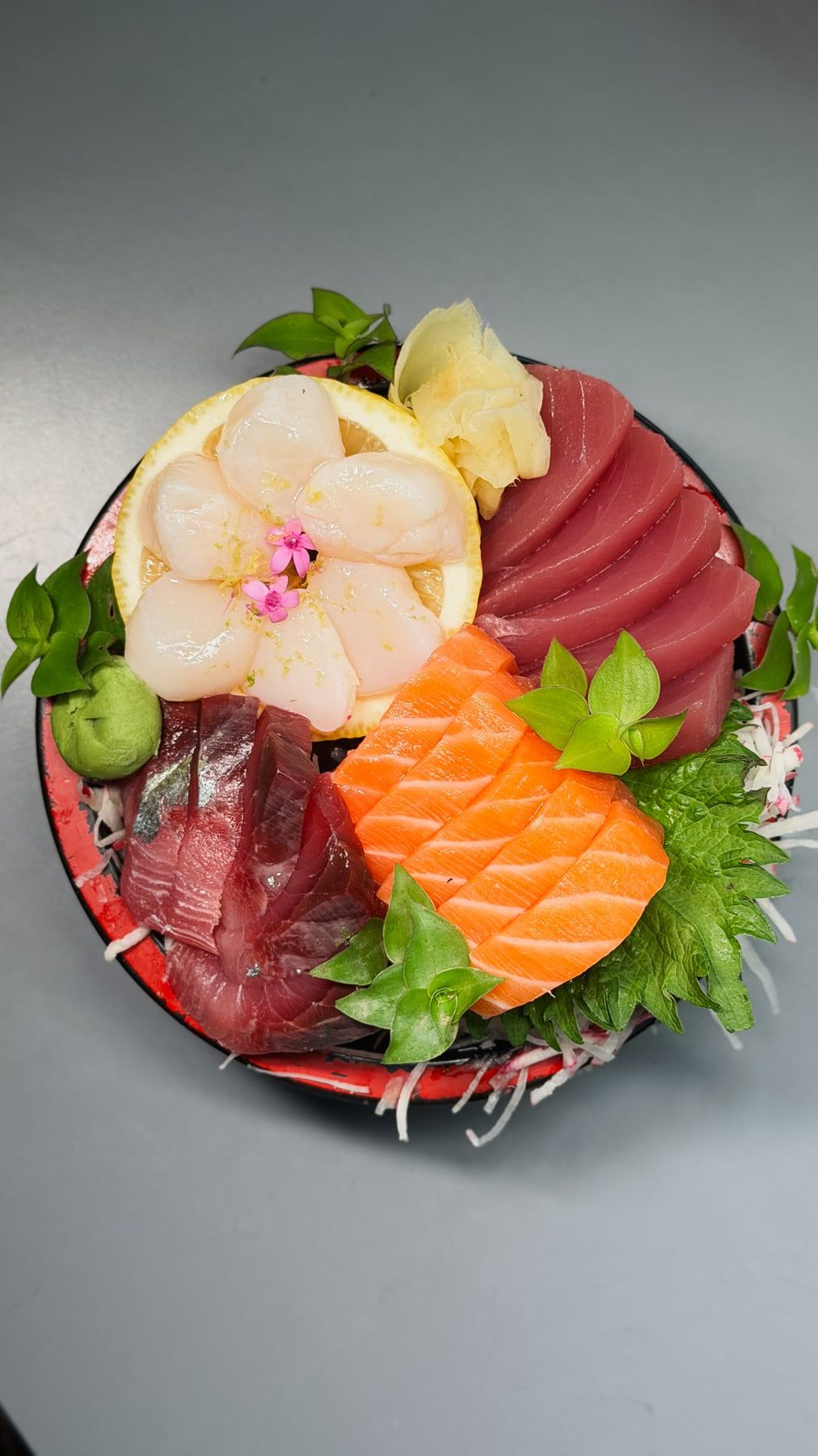 Travessa de sashimis vista de cima: atum, salmão e vieiras com flores, gengibre e wasabi sobre folhas de shissô.