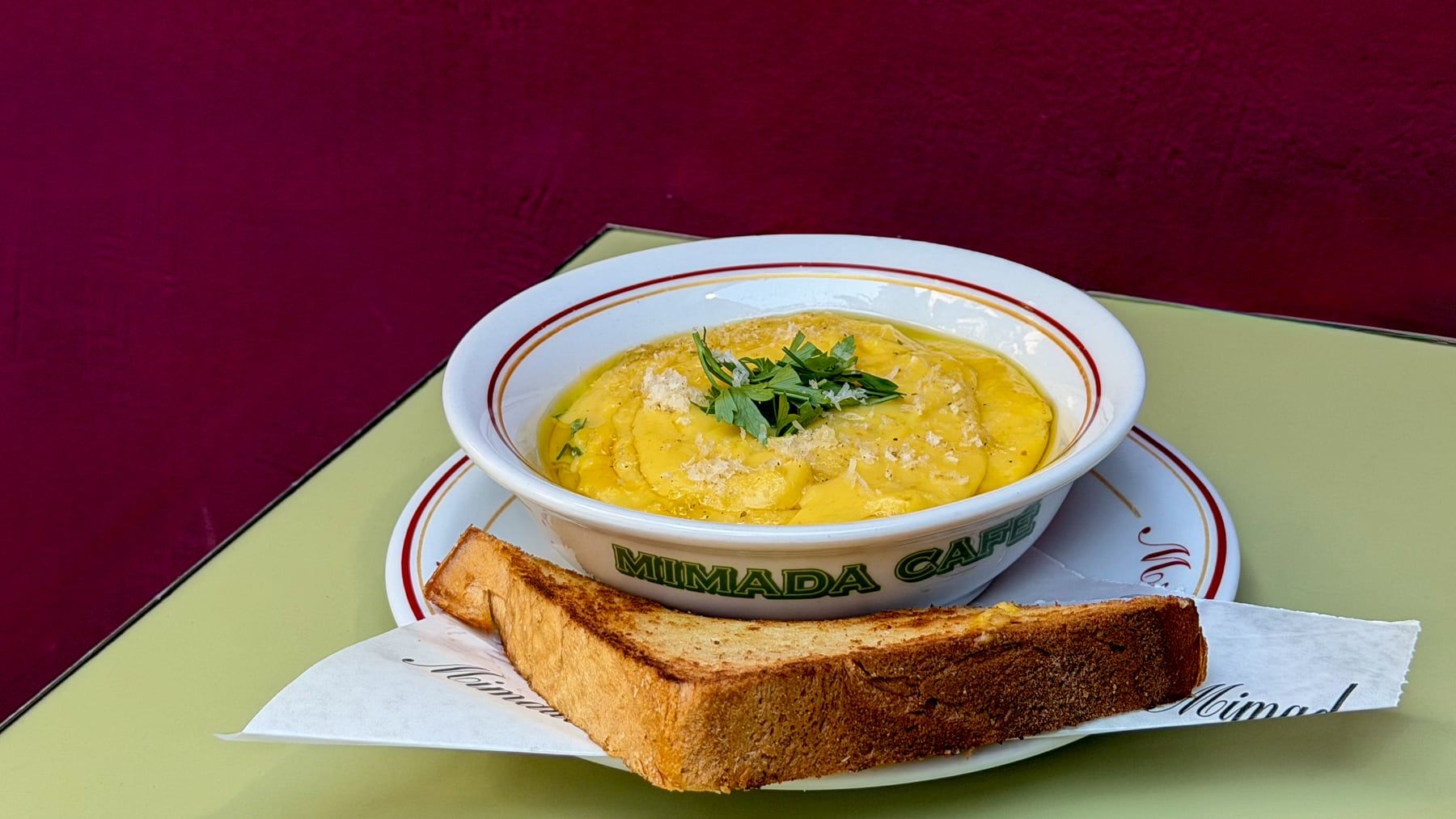 Tigela com a inscrição Mimada Café, com um creme amarelo e ervas, e fatias de pão tostado ao lado.