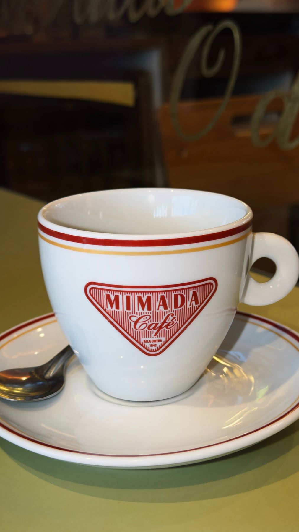 Xícara branca de borda vermelha com o losango do Mimada Café estampado, sobre o pires.